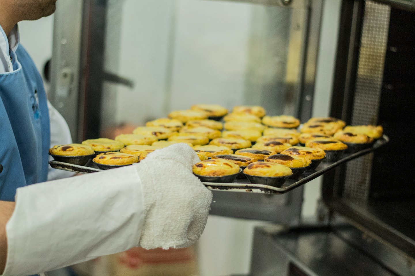 Baker in a blue apron sliding egg tarts into an oven.